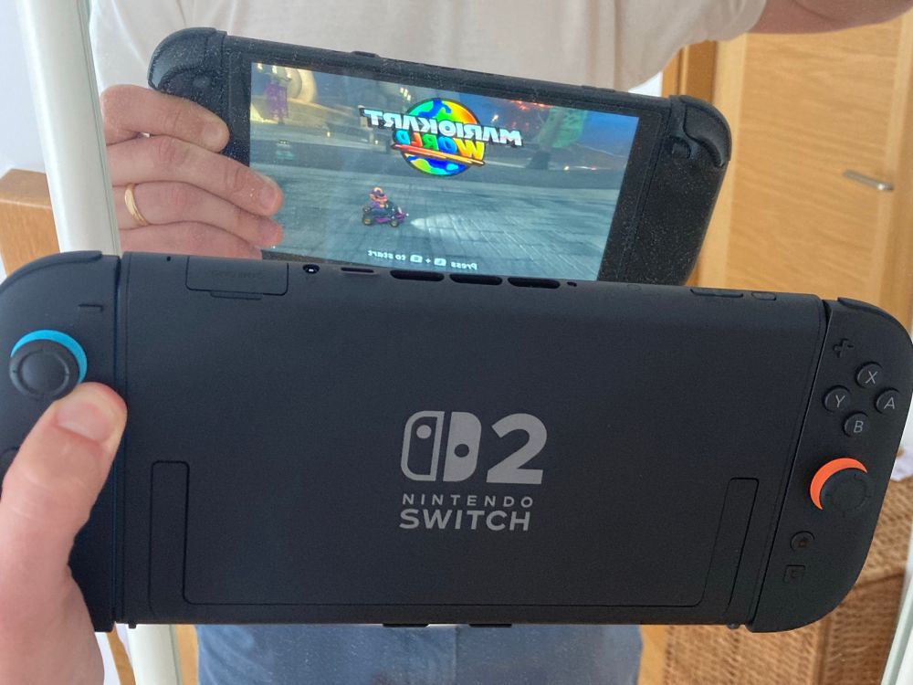 A man in a white t-shirt holding a Switch 2 console in front of a mirror with Joy-Con controllers attached back-to-front.

Mario Kart World’s menu screen is seen on the screen in the reflection.