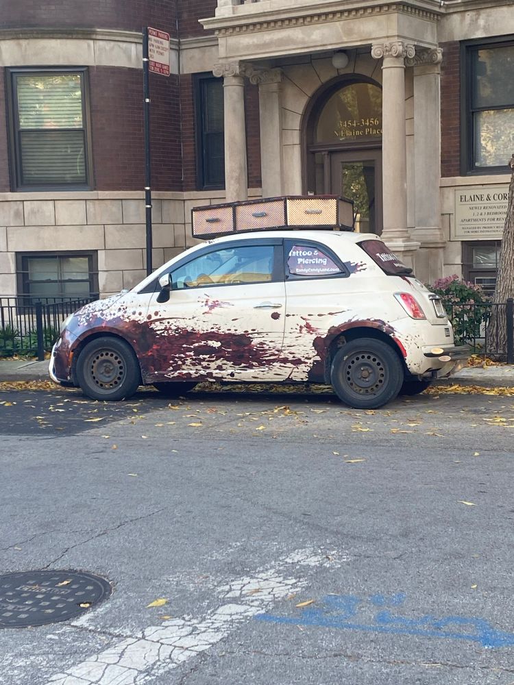 A small white car with splattered blood stains all over the front quarter panel. There’s a tattered sign in the window that says tattoo and piercing. there appears to be a coffin strap to the roof.