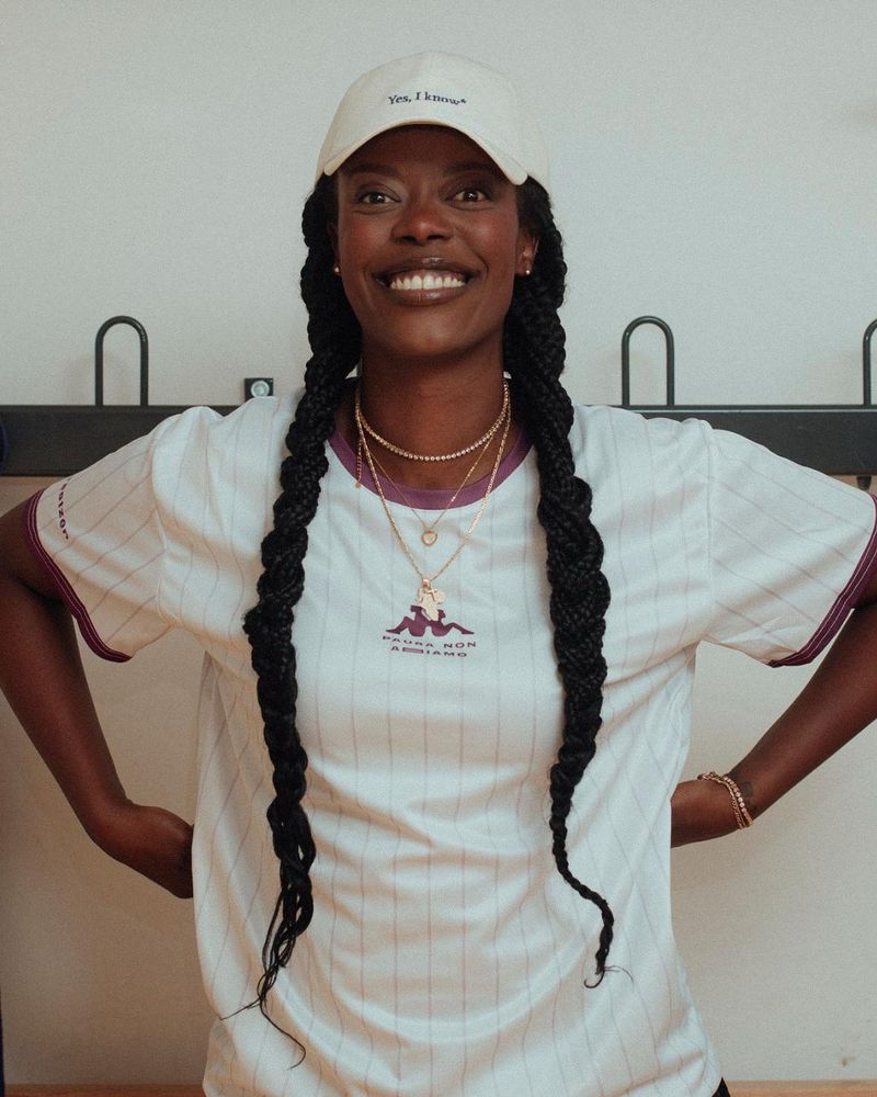 A smiling person with two long braids and a white cap wears a white shirt with purple details and looks directly into the camera.