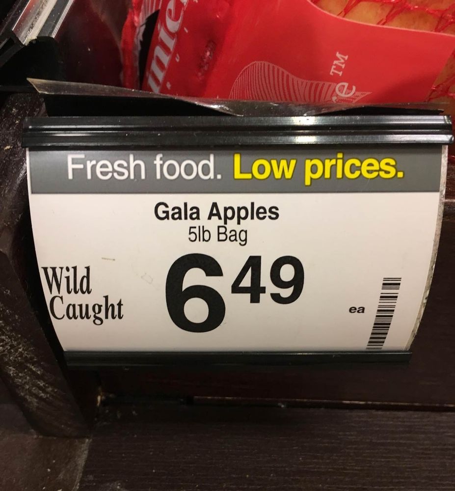 A close-up shot of a price tag at a grocery store shelf displays the details for Gala apples. The sign announces "Fresh food. Low prices." with the product information below, listing "Gala Apples 5lb Bag" and a price of "6.49" with "ea" to the side. The tag also includes the words "Wild Caught." A blurry image of the bagged apples sits behind the sign