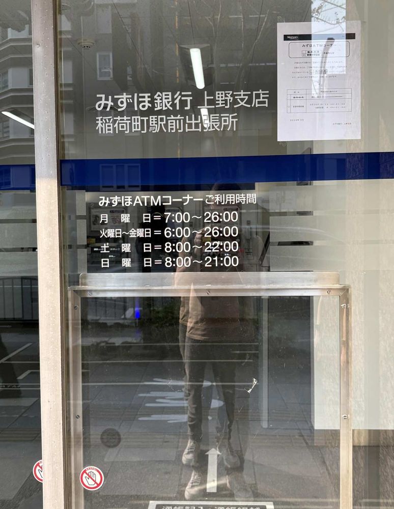 A storefront with information on Mizuho Bank, Ueno Branch, including the bank name written in Japanese, details for Inaricho Station, and ATM hours of operation in black lettering on a transparent board. Hours are 7 AM-10 PM weekdays, 8-22 Saturday, and 8-21 Sunday. A notice about time changes on March 30th is posted.