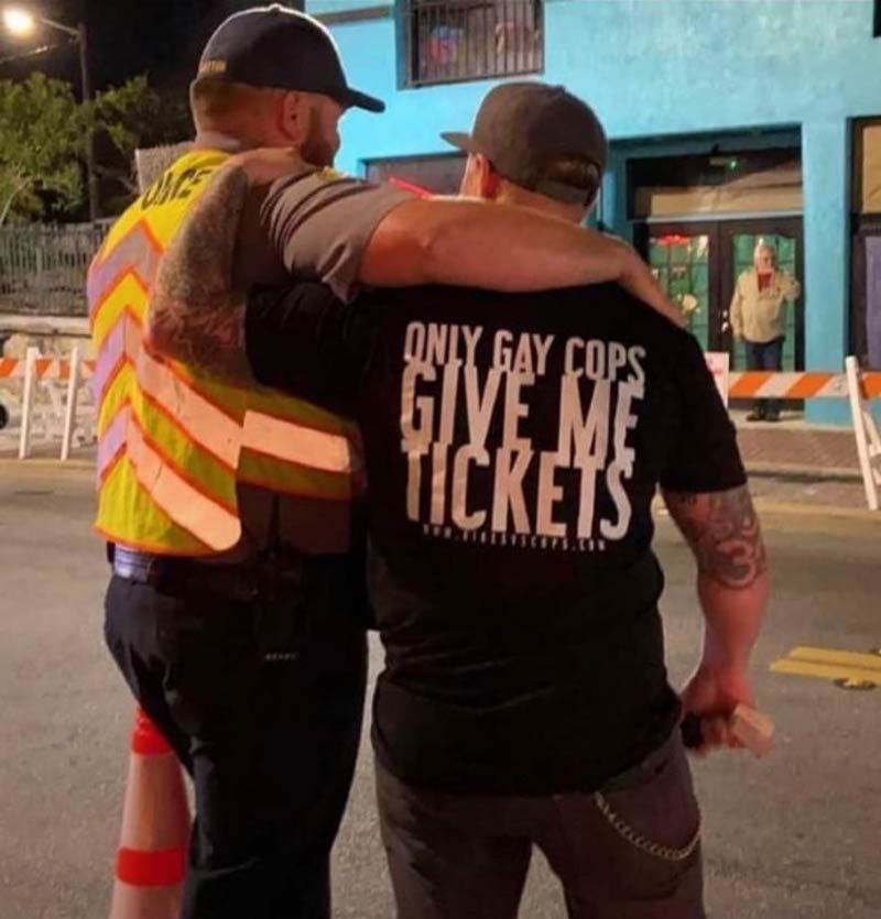 Two men, one wearing a traffic vest and the other a t-shirt that says "Only Gay Cops Give Me Tickets," stand arm in arm.