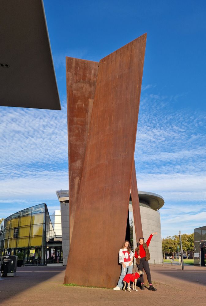 Marie Lotte, Nydia en hond Toby poseren (alle drie in het rood) bij een 17 meter hoog kunstwerk dat bestaat uit roestkleurige stalen platen die tegen elkaar aan staan
