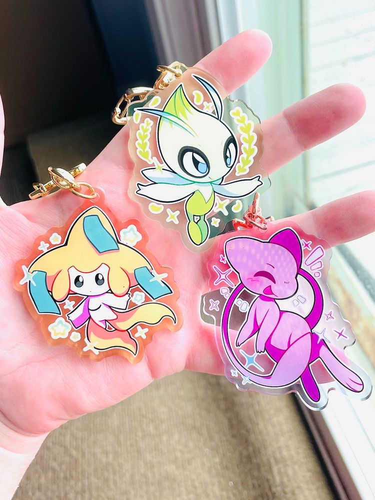 a photo of a hand holding up three keychains. the keychains display celebi, jirachi, and mew from pokemon.