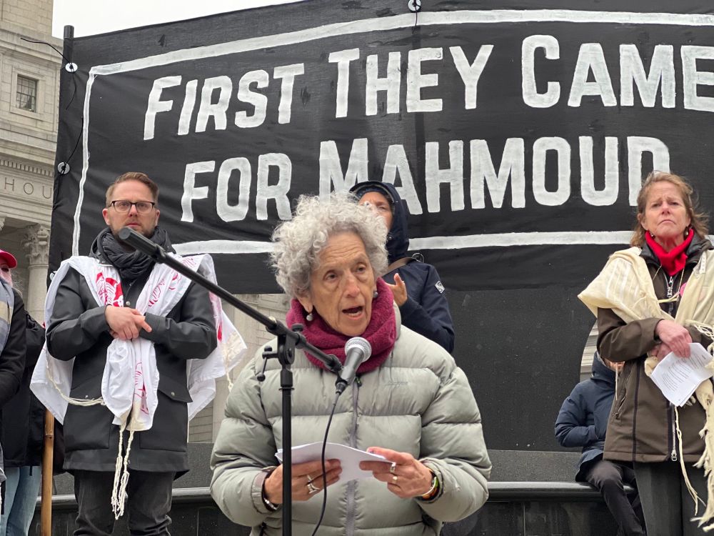 an elderly woman speaking at a mic, behind her are three other people looking solemn, and a banner that reads "First They Came For Mahmoud"