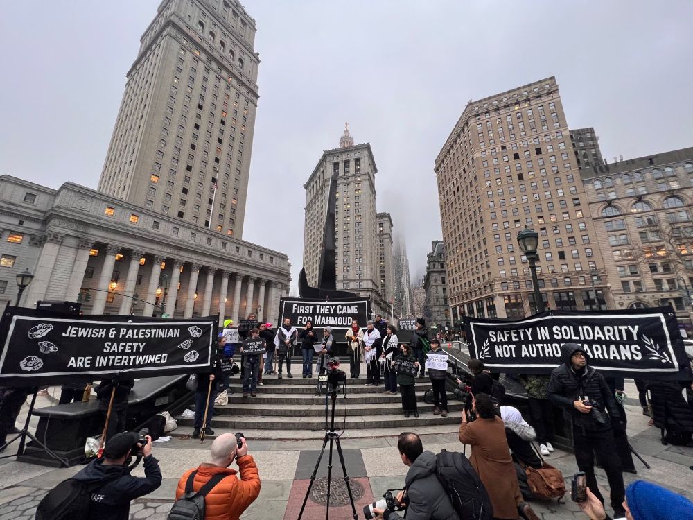 speakers gathered on steps in the city square. banners with white on black text read: "Jewish & Palestinian Safety Are Intertwined", "First They Came For Mahmoud", and "Safety In Solidarity, Not Authoritarians". In the foreground, we see press photographers from the back, facing the speakers.