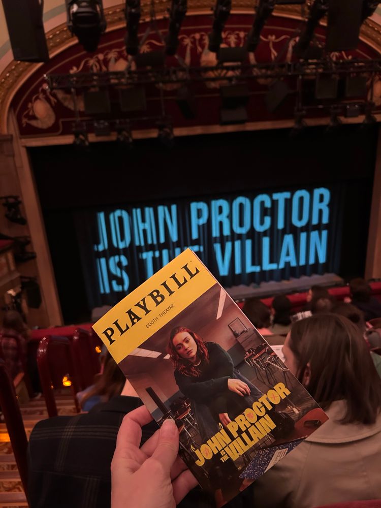 A hand holds a Playbill in the mezzanine of the Booth Theatre in NYC. Sadie Sink in a green sweater sits on a desk in a high school classroom in the image on the Playbill. Text on both the Playbill and the projected on the curtain is JOHN PROCTOR IS THE VILLAIN. The play is by Kimberly Belflower