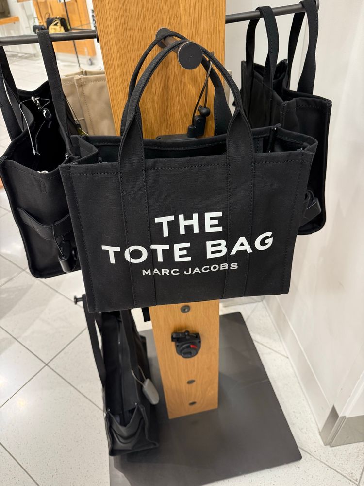 A tote bag on a rack at a store. On the side in large letters is printed THE TOTE BAG and the name of designer Marc Jacobs