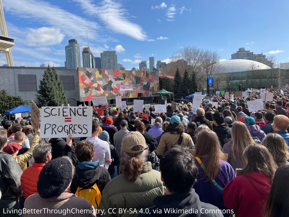 A crowd of people at a protest. One is holding a sign that reads “science = progress”
