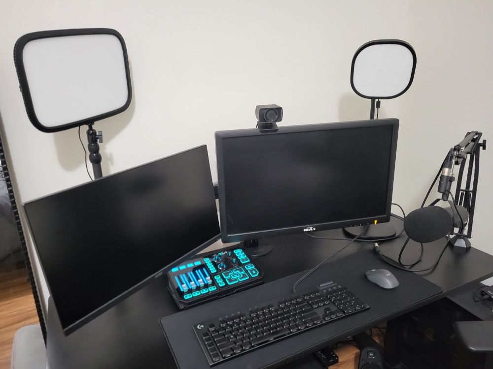 A photo of a streaming setup - 
A GoXLR, two monitors on adjustable arms, a mounted Shure SM7B mic, two Elgato lights, a plain black mat, a mouse, and a keyboard are organized neatly on a black desk. A small Elgato webcam perches on top of a monitor.