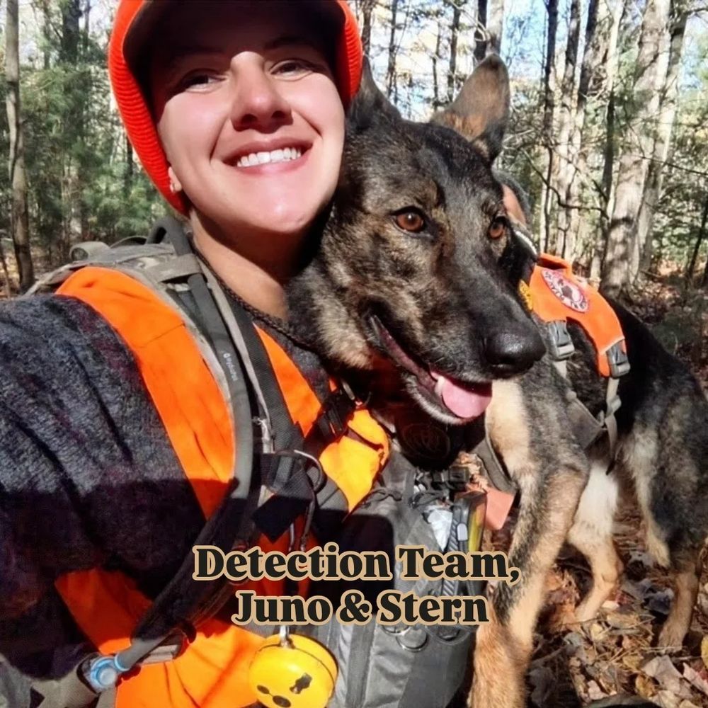 A woman in an orange hat smiles at the camera with her detection dog, a Shepard mix, next to her face. They are outside.