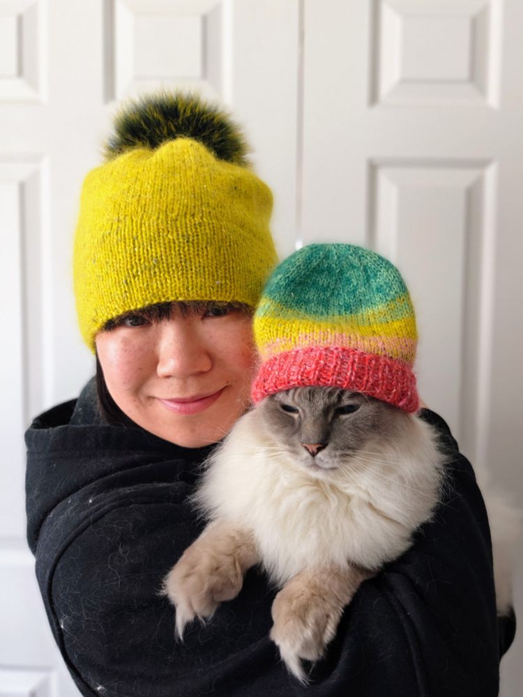 Closeup portrait photo of an Asian woman holding a ragdoll cat. Both are wearing fluffy knitted hats in bright colours