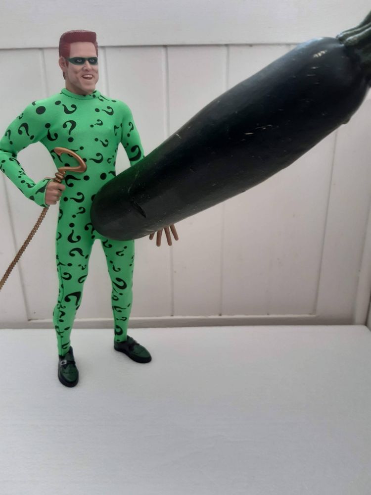 A Jim Carrey Riddler doll posed with a large courgette. His face is grinning and the vegetable is extending out from his crotch in a suggestive manner.