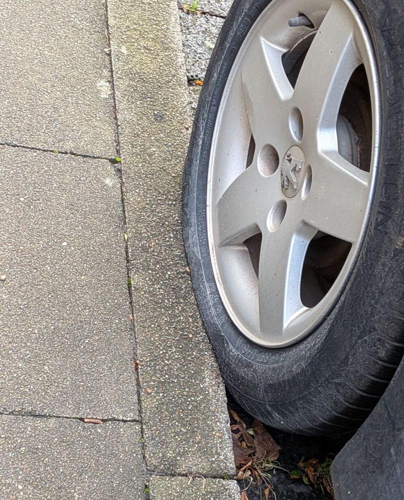 Der Reifen eines geparkten Autos. Der Reifen parkt zum Teil auf dem Gehweg, sodass sich der Gehwegrand quasi in den Reifen hineinbohrt und ihn sehr ungesund verformt. 