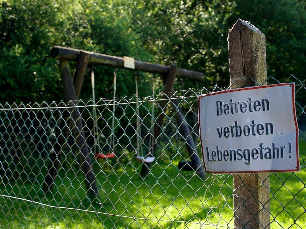 Im Vordergrund ein Zaun mit einem Schild "Betreten verboten, Lebensgefahr!" 

Im Hintergrund ein Kinderspielplatz mit Schaukeln. 