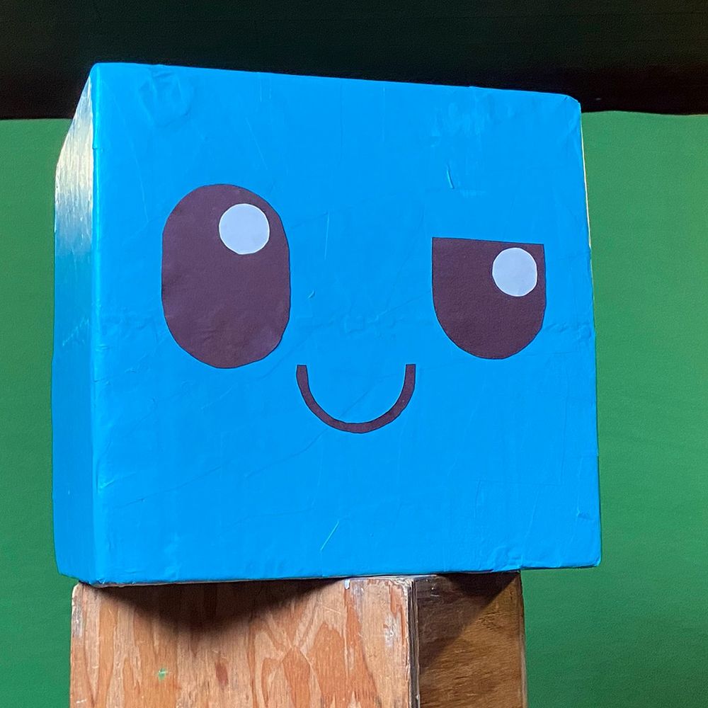 A recreation of Cold Fusion Comedy's ice cube mascot logo. Built out of styrofoam, paper mache, construction paper, glue, and top coat. A blue cube with two eyes cartoon looking in the upper right corner and a smile. It rests on top of a wooden apple box in front of a green screen.