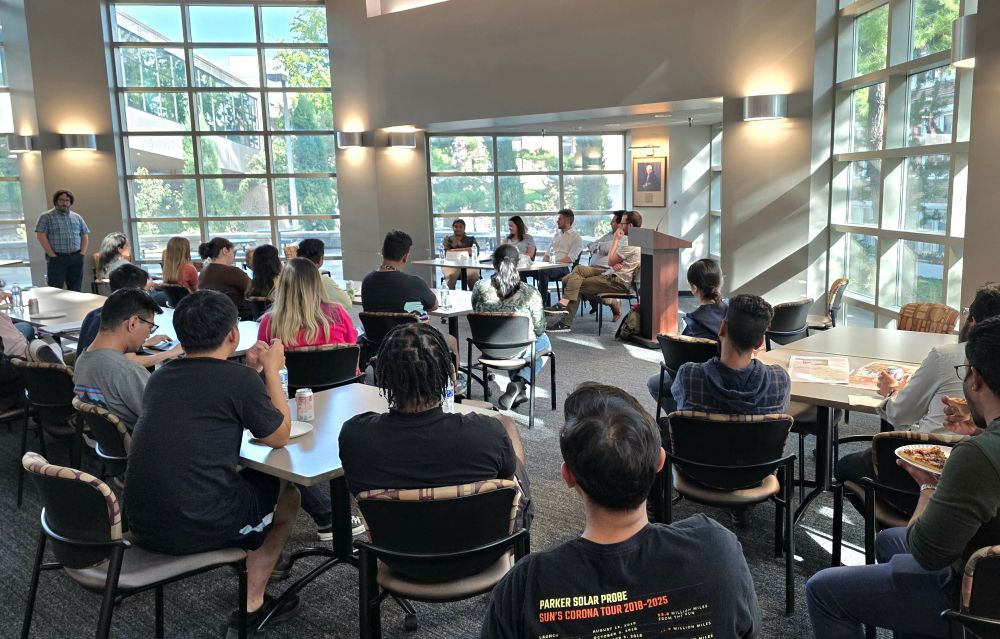 Postdocs seated in a sunlit room, facing a panel of five professors seated at a table in the front of the room, who are in front of floor-to-ceiling windows.
