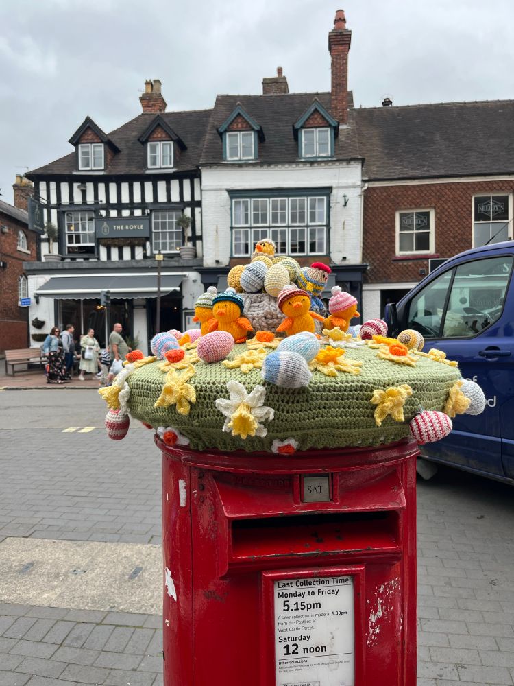 Red post box with crocheted topper of daffodils, eggs and chicks. Behind, the pavement and a black and white timber building. The sky is overcast, cloudy and grey. 