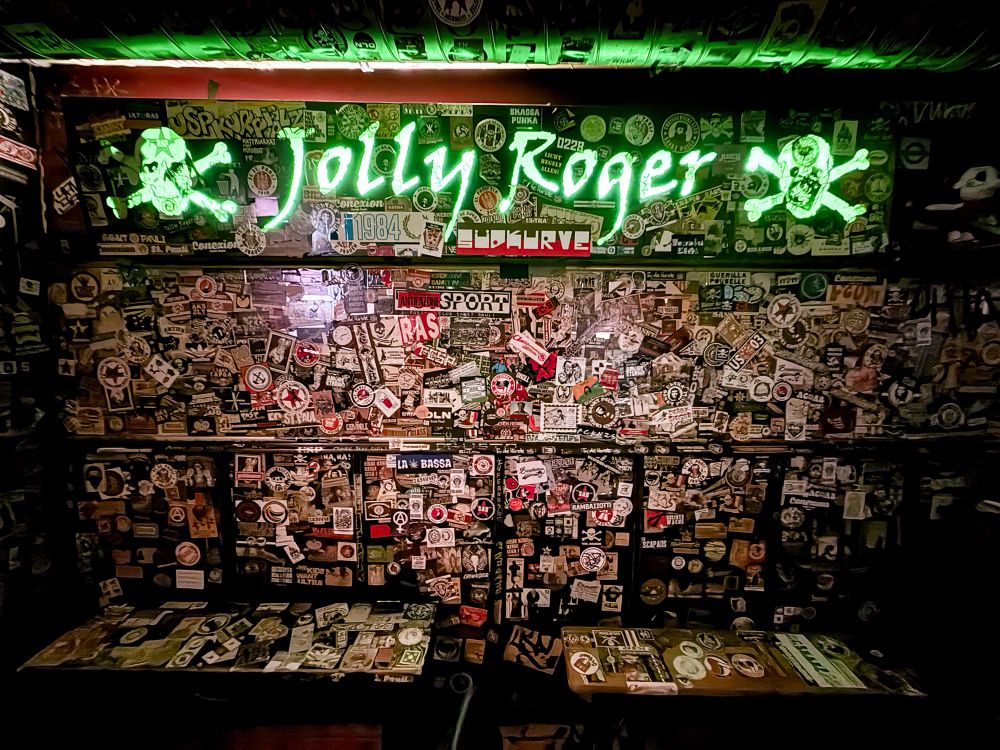 Photo of the back wall of the Jolly Roger bar in St Pauli, Hamburg. The entire wall is covered in anti-fascist stickers, and there’s a green neon Jolly Roger sign flanked by two green neon totenkopfs. 