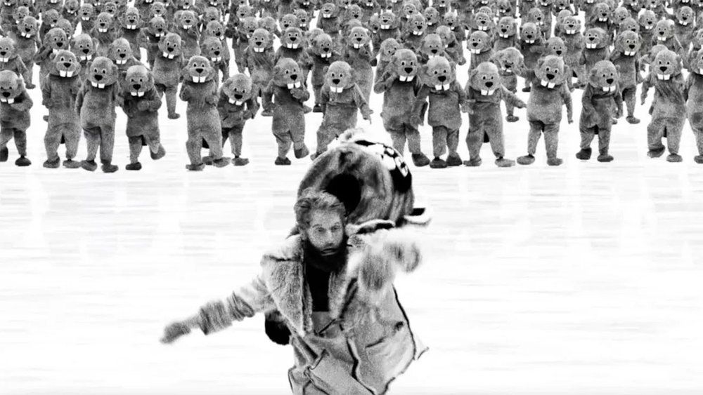 Bilde fra «Hundreds of Beavers» i svart-hvit: En mann med skjegg og kledd i pels løper desperat mot kameraet. Bak ham kommer en stor folkmengde av folk i tegneserieaktige beverkostymer.