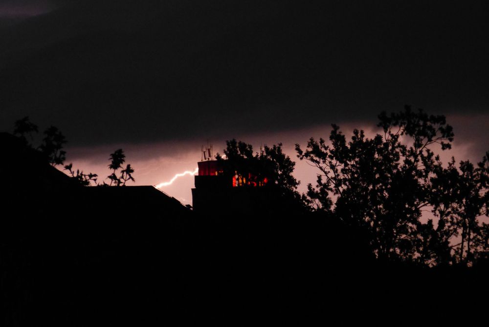 A photo taken at night during a thunderstorm. The black silhouette of house roofs and trees stand out against a lightning-lit overcast sky. The prominent silhouette of a tower with red-lit window arches stands out the most, it almost looks like the lightning is cast from one of those windows.