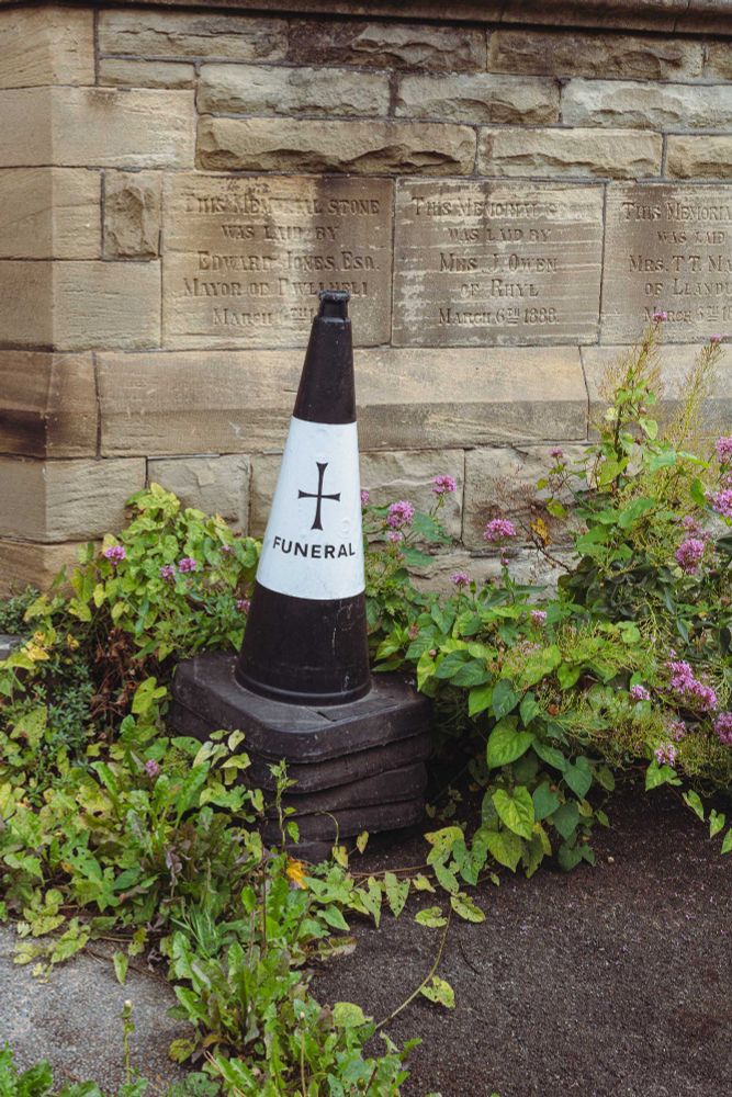 This photo shows a black and white traffic cone marked with a cross and the word "FUNERAL", placed on a stack of square black bases. The cone is situated in a small garden bed surrounded by overgrown greenery and purple flowers. Behind the cone is a sandstone wall with multiple memorial plaques carved into it. The plaques mention various names and dates, such as Edward Jones, Esq., Mayor of Llandudno, and dates like March 6th, 1888, and March 9th, 1888.
The juxtaposition of the sombre funeral cone with the slightly unkempt garden and old memorials gives the scene a quietly surreal or slightly humorous tone. It appears to be outside a church or chapel, possibly marking reserved parking or access for a funeral service.