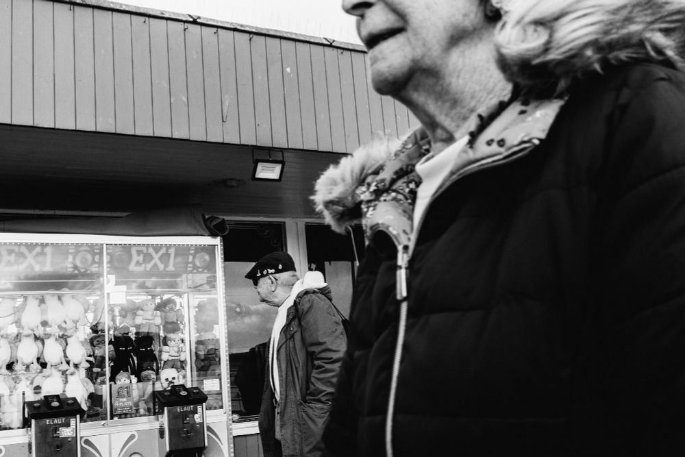 Black and white street photograph showing an older man in a flat cap and coat standing outside a seaside arcade filled with plush toys, while a woman in a hooded jacket passes close to the camera, partially obscuring the frame.
