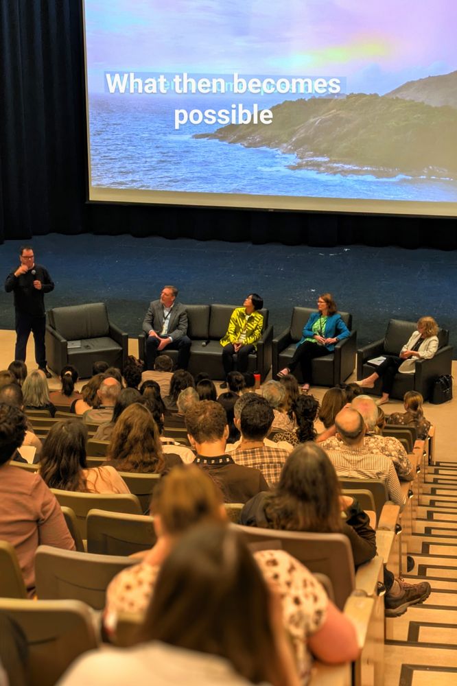 Five panelists sitting at the front of an auditorium with an audience watching them. A slide behind says what then becomes possible with a picture of a coast. 