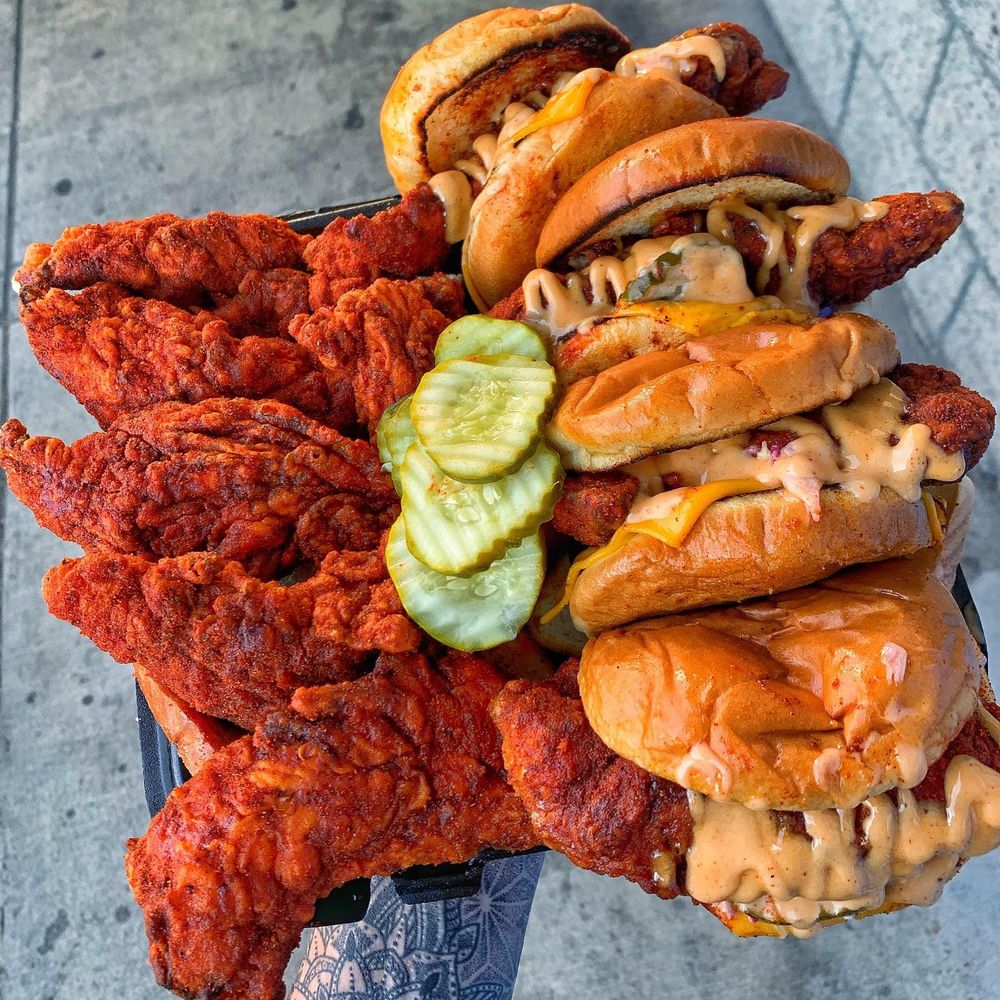 A close-up, overhead shot of a black tray overflowing with food. The left side is piled high with bright red, crispy fried chicken tenders, while the right side features a stack of several chicken sandwiches on toasted buns, dripping with melted cheese and a creamy orange sauce. A pile of dill pickle slices rests in the center. The tray is held by a tattooed hand over a gray concrete background.