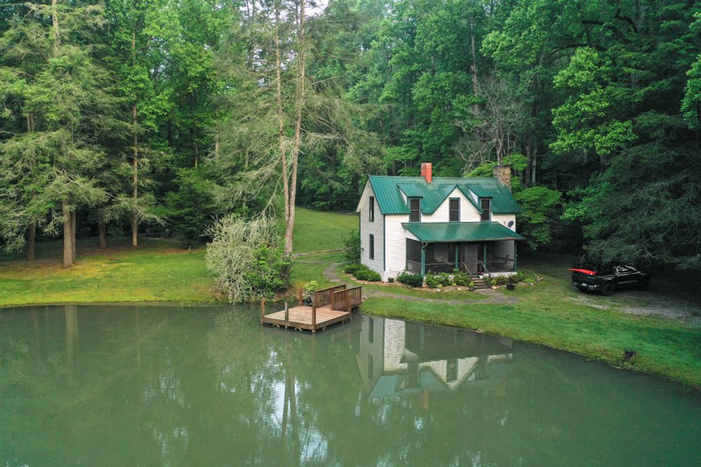 A tranquil aerial shot of a small white house with a green metal roof and a red brick chimney. The house, named Big Creek Lodge, sits on a grassy lawn next to a calm pond with a small wooden deck extending over the water. A black pickup truck is parked on a dirt road to the right of the house. The entire scene is surrounded by a dense green forest under a cloudy sky. The reflection of the house is visible in the still water of the pond.
