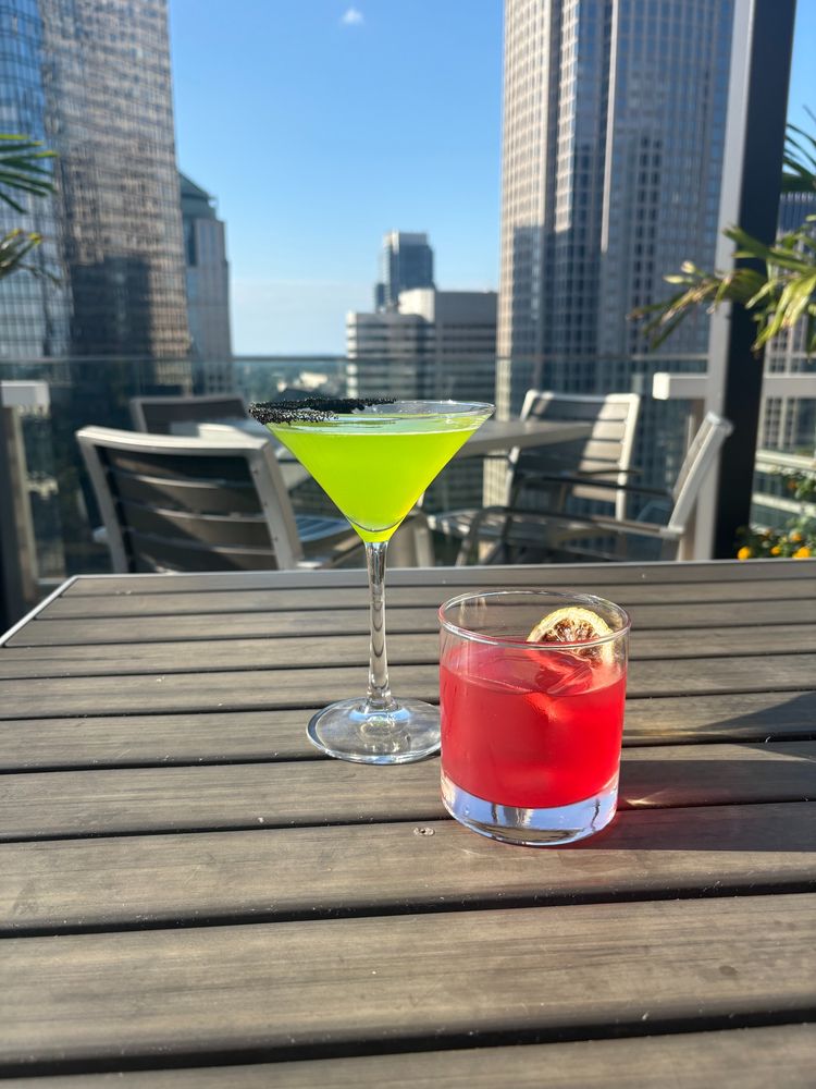 A close-up of two vibrant cocktails on an outdoor rooftop deck. The drink in the back is a green martini in a glass with a black sugar rim. In the front is a red drink on the rocks, garnished with a citrus wheel. Tall city buildings and a clear blue sky are visible in the background.