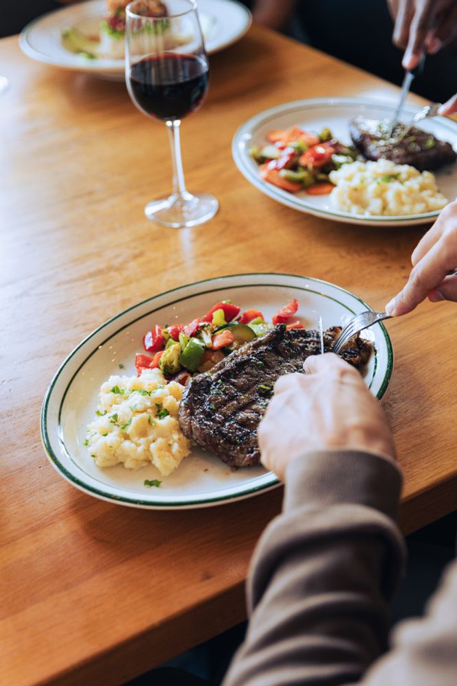 A person uses a knife and fork to cut into a grilled steak on a plate. The steak is served with mashed potatoes and mixed vegetables.