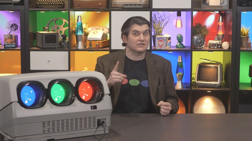 A YouTuber in a brown jacket behind a desk points next to a 3-CRT video projector on the desk that matches the look of his t-shirt (with the colors in a different order).
The background is a bookshelf of square cubbies with 3 lava lamps (1 red, 1 green, 1 blue) amongst various other items in front of a block of solid color, different in each cubbie.