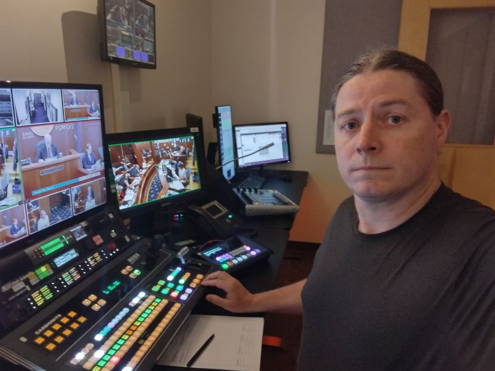 A person in a black t-shirt sitting in a TV control room. There are monitors on the left and control panels on the table in the middle.