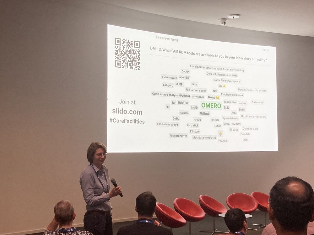 Photo of Stefanie Weidtkamp-Peters standing in front of a Slido presentation. There are many smaller answers to the question including "idk", "🤷", "USB stick", and similar but one large green word: "OMERO"