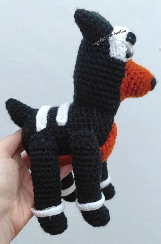 Houndour amigurumi, viewed from the side. In black, copper and white. The Houndour has two white straps on its back, a white strap in every leg, and a skull mask also in white. I tried to recreate the "anime eyes", but they blend with the white mask. 