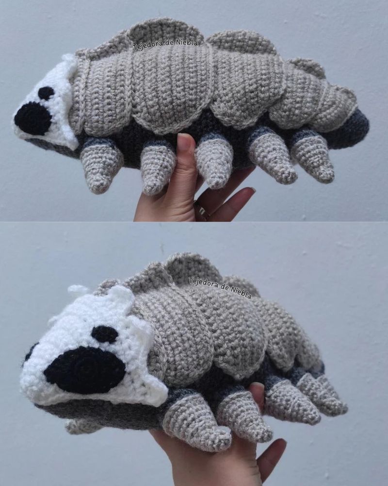Amigurumi Bell Beast, from Silksong. It's 2 different pictures, the top one shows the beast from the side, and the bottom one it's a bit tilted to show the mask better. In dark and light grey, black and white. The beast has 10 legs and 6 "scales" in the back. 