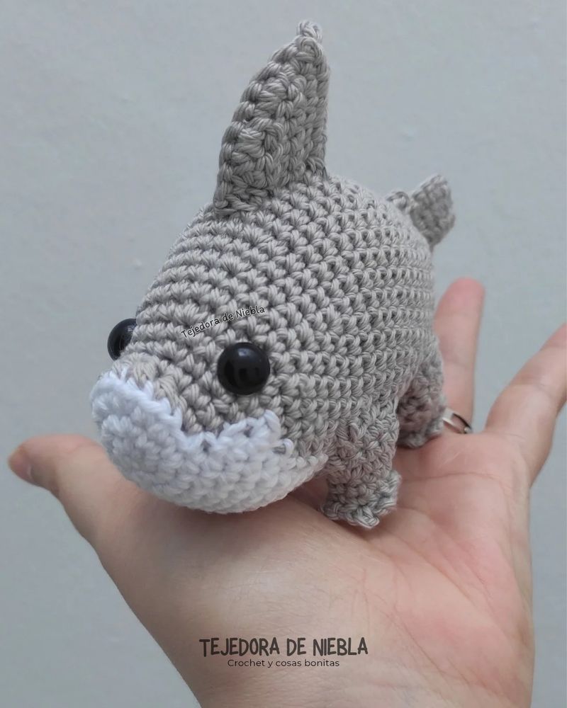Amigurumi de un tiburón de color gris y blanco, con cuatro patas. Está apoyado sobre la palma de mi mano. 