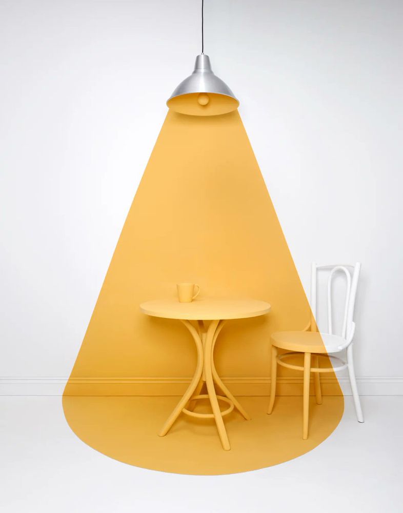 The image features a cleverly designed scene where a silver pendant lamp appears to cast a yellow cone of light, but the "light" is actually painted on the table, chair, and wall. creating an illusion of illumination. A yellow table with a matching cup sits within the painted area, while the white chair is only partially painted to match the effect. https://alexander-kent.com/ 