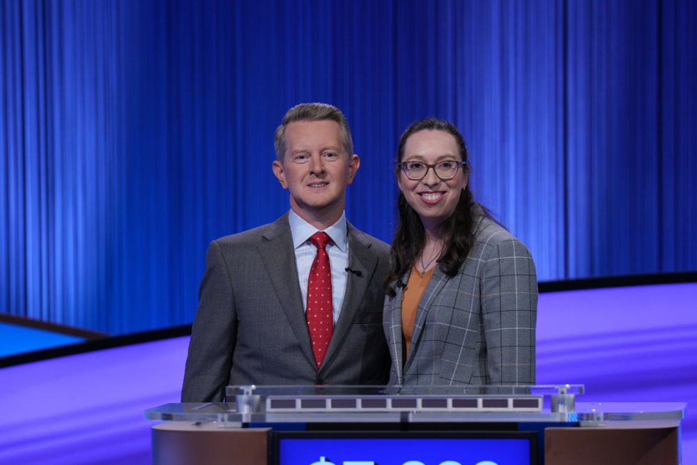 Ken Jennings in a charcoal suit and red tie, next to me, a white woman with glasses and dark brown hair, a yellow-orange shirt, a gray blazer with checkerboard striping, and a huge smile. We're standing at the real Jeopardy! podium with the show's blue and purple set in the background