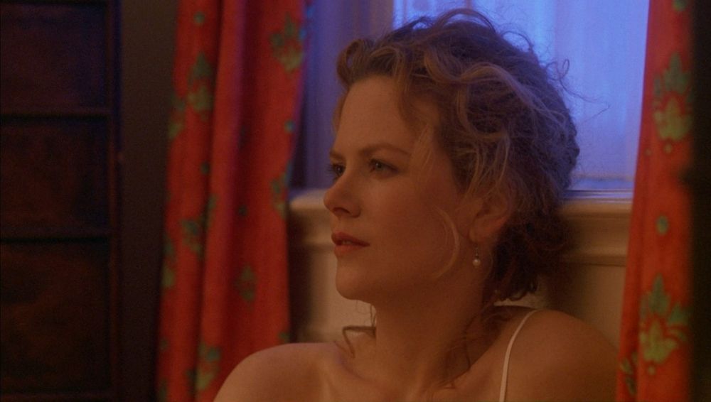 A medium close-up of Alice, sitting beside the window, looking at Bill; red curtains with leafy green patterns to either side of her; blue light from the window behind her head.