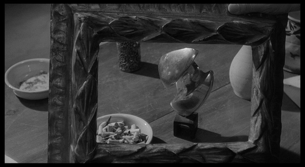 From Antonioni's L'eclisse: a close-up of an empty picture frame through which objects can be seen on a table (an abstract sculpture, an ashtray full of cigarette buts, a gourd, a cup or candle-holder); in the upper right, we can see the hand of Vittoria (Monica Vitti) holding the frame in place.