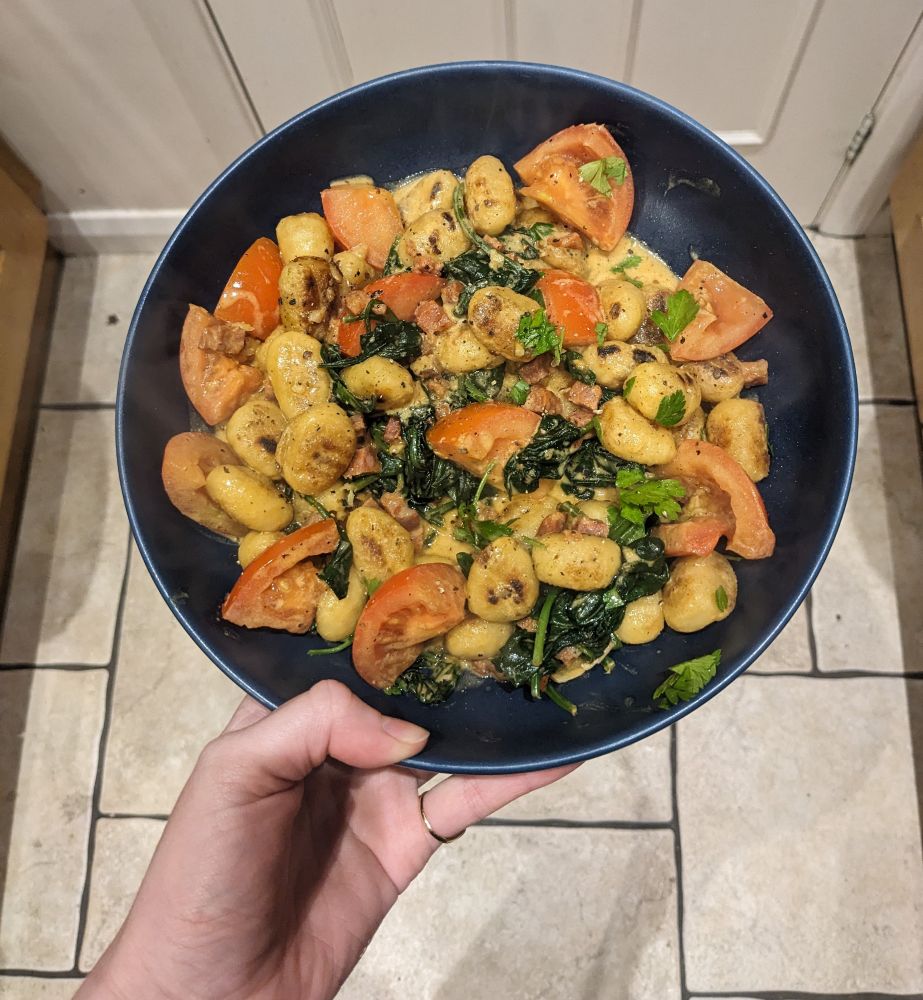 A white hand holding a blue bowl. In it is a dish of gnocchi, tomato, spinach, and parsley with a chorizo crème fraiche sauce. 