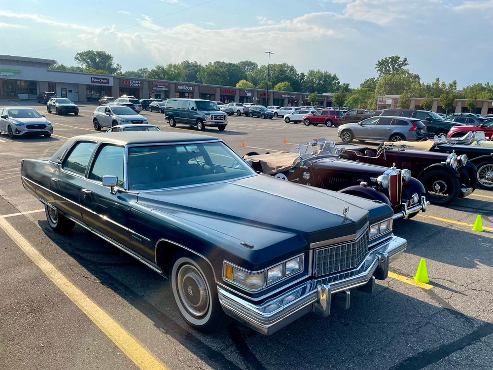 Photograph in a parking lot of a dark blue mid-70s Cadillac Fleetwood Brougham seen from the front right with a small 1952 MG TD next to and behind it, and several other small MG T-Series cars behind the TD.