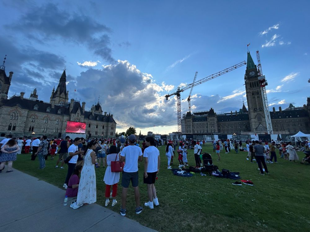 ottawa’s parliament hill on a cloudy Canada Day. there’s a moderate crowd standing around facing a small screen on the left side of the hill. in the background, the main tower of parliament rises alongside several construction cranes.