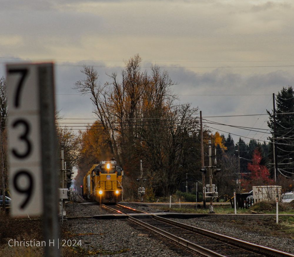 In the foreground, a vertical white sign with black lettering reads 739, indicating the railroad milepost. Beyond it, a yellow Union Pacific locomotive is in the process of crossing one grade crossing and approaching another. We can see it pulling a few cars. Trees with no leaves and trees with orange and red leaves line the tracks.