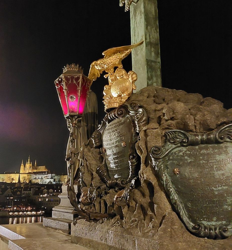 Sockel der Skulptur des Kalvarienbergs, die Lampe mit dem Adler leuchtet rot vor dem Hintergrund der beleuchteten Stadt jenseits der Brücke.