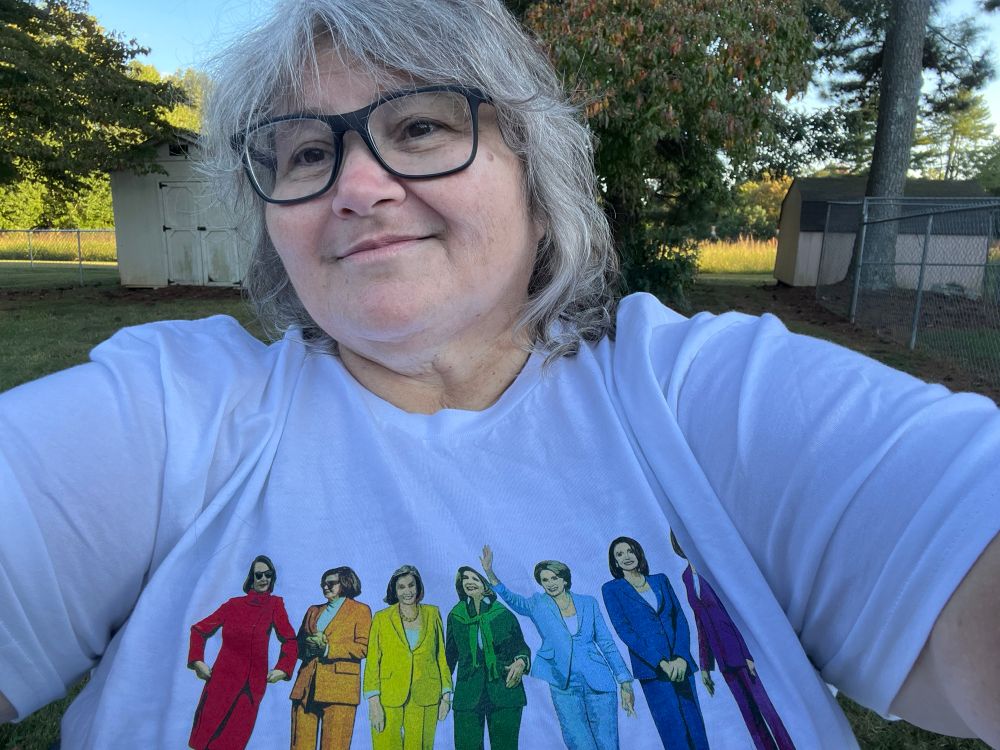 Pamela (grey haired woman with glasses) wearing a white T shirt with multiposed Nancy Pelosi in suits that create a rainbow 🌈 of Nancy Pelosis 