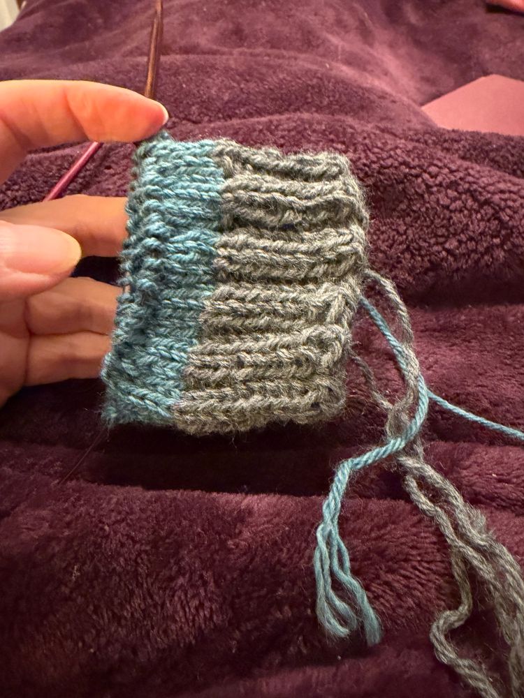 The beginning of a worsted weight hand knit sock with grey 2x2 ribbing cuff and blue 3x1 ribbing body. 