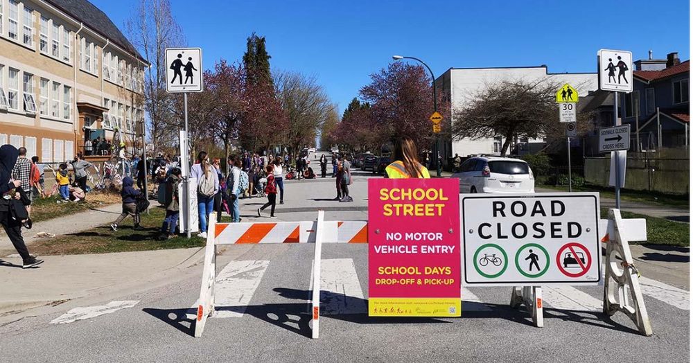 A street is open to kids and families for 30 minutes before school. In the foreground is a barricade closing it to cars, kids make their way to school on a stress free walk.